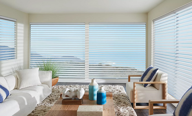 Sunlight enters through Hunter Douglas's blinds, illuminating a white round table with four white chairs. On the table is a plate of oranges and a vase of yellow flowers.