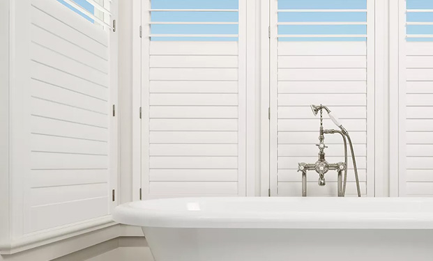 In a serene bathroom setting, sunlight filters through moisture-resistant Hunter Douglas window blinds, casting gentle rays onto a pristine white bathtub, faucet, and shower.